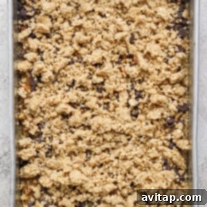 A rectangular baking tray filled with an unbaked dessert mixture, elegantly crowned with a delightful crumbly oat and nut topping, positioned on a textured light-colored surface.