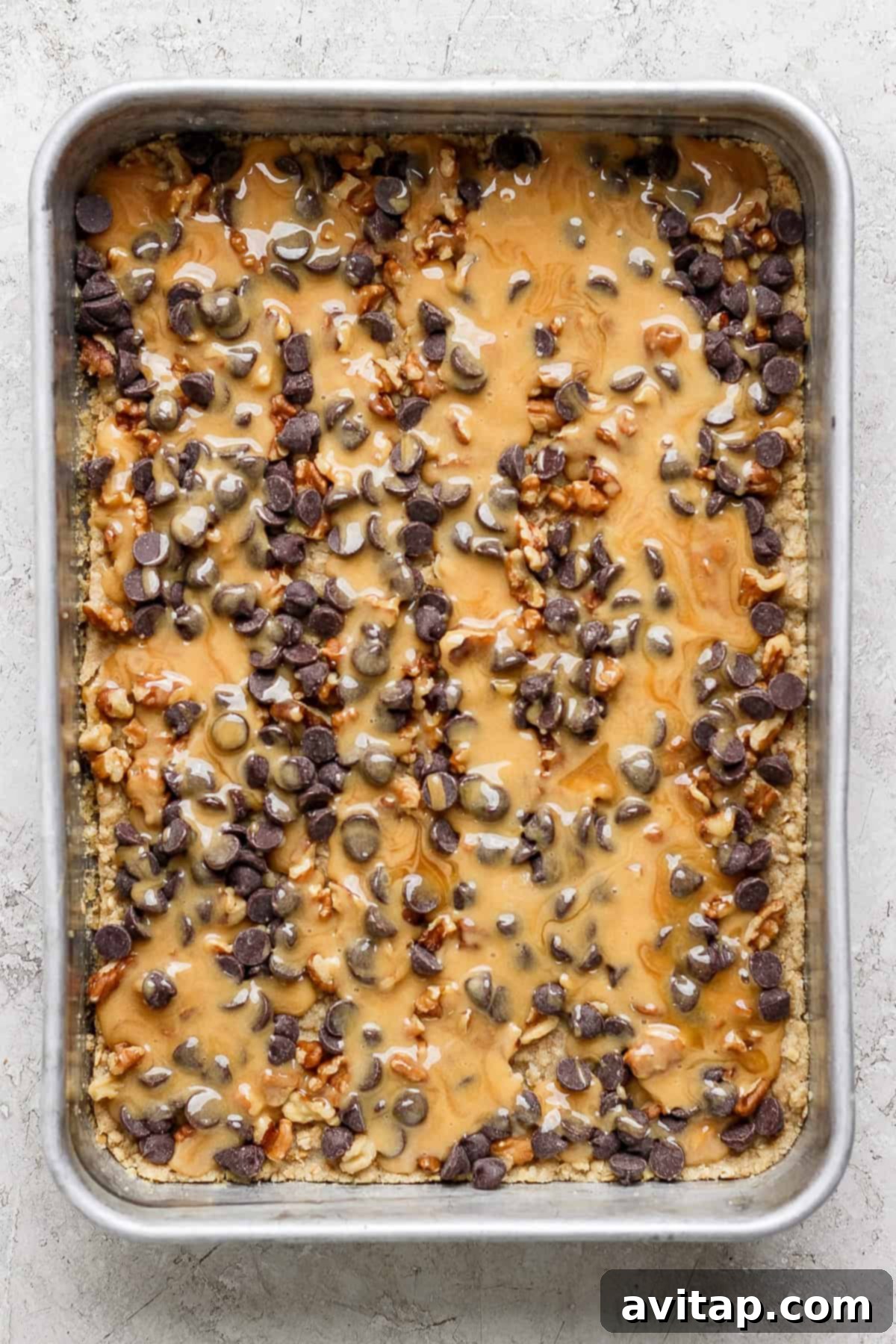 A baking tray filled with a delicious dessert topped with glistening melted caramel, generous chocolate chips, and finely chopped nuts, ready for its final bake.