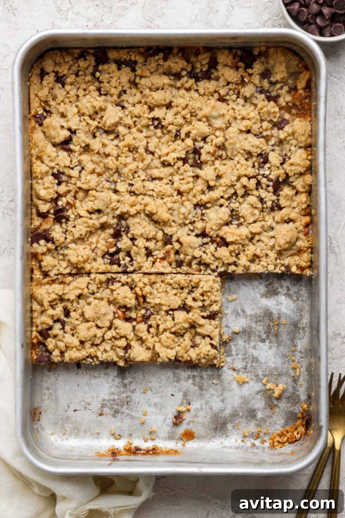 A metal baking pan filled with golden-brown crumbly dessert bars. Two bars have been neatly cut out, showcasing their luscious, layered filling of caramel and chocolate. A fork and a pristine white napkin rest nearby, inviting a taste.