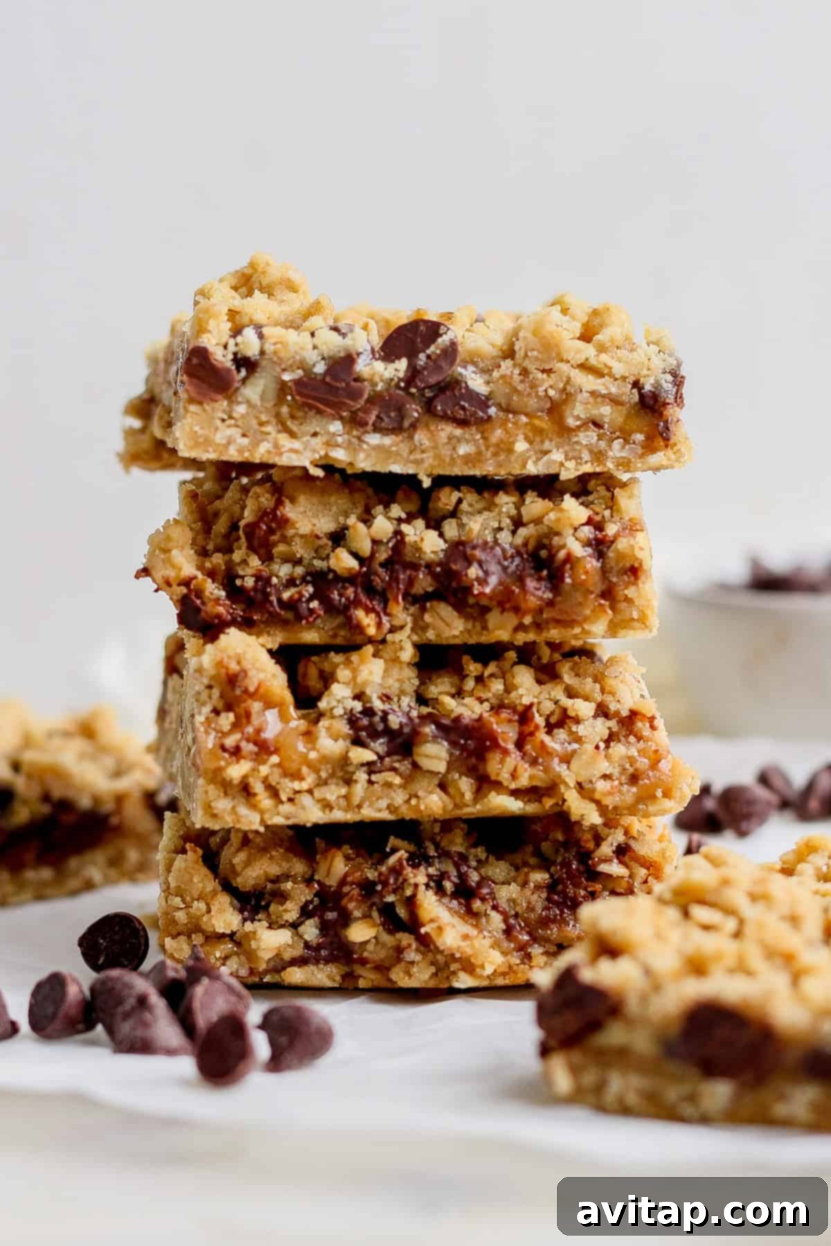 A perfect stack of four chocolate chip oatmeal bars, each featuring a delightful crumbly top, is artfully displayed on a pristine white surface. A scattering of chocolate chips surrounds the stack, enhancing the visual appeal.