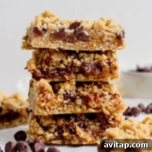 A stack of four delicious chocolate chip oatmeal bars with a delightful crumbly texture, presented on a clean white surface and surrounded by scattered chocolate chips, ready to be enjoyed.