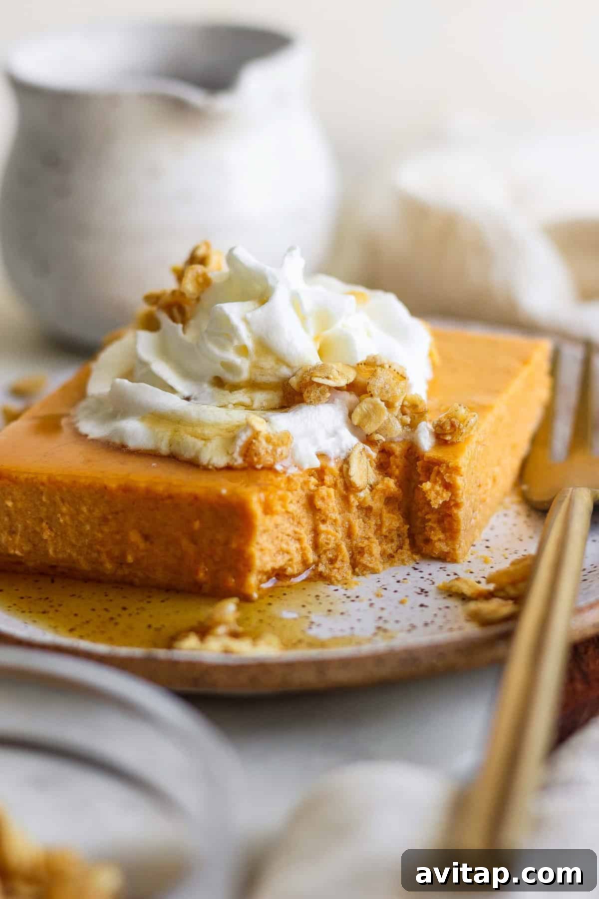 A perfectly sliced high-protein pumpkin bar, topped generously with creamy whipped cream, crunchy granola, and a sweet drizzle of maple syrup, resting on a rustic plate beside a fork, inviting you to take a bite.