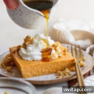 A hand drizzles rich maple syrup from a small jug onto a delectable slice of cheesecake, already topped with a cloud of whipped cream and a sprinkle of granola, beautifully presented on a plate next to a fork.