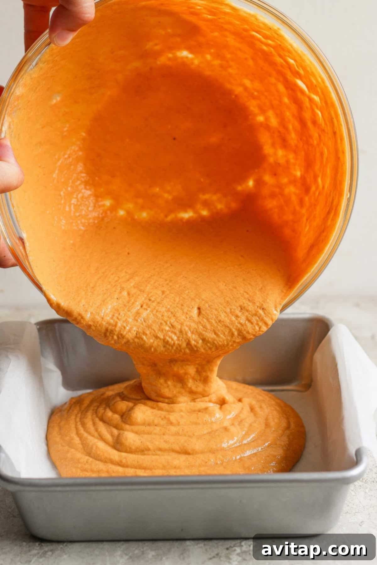 A pair of hands carefully pouring thick, orange-colored batter from a large mixing bowl into a parchment-lined square baking tin, preparing it for the oven.