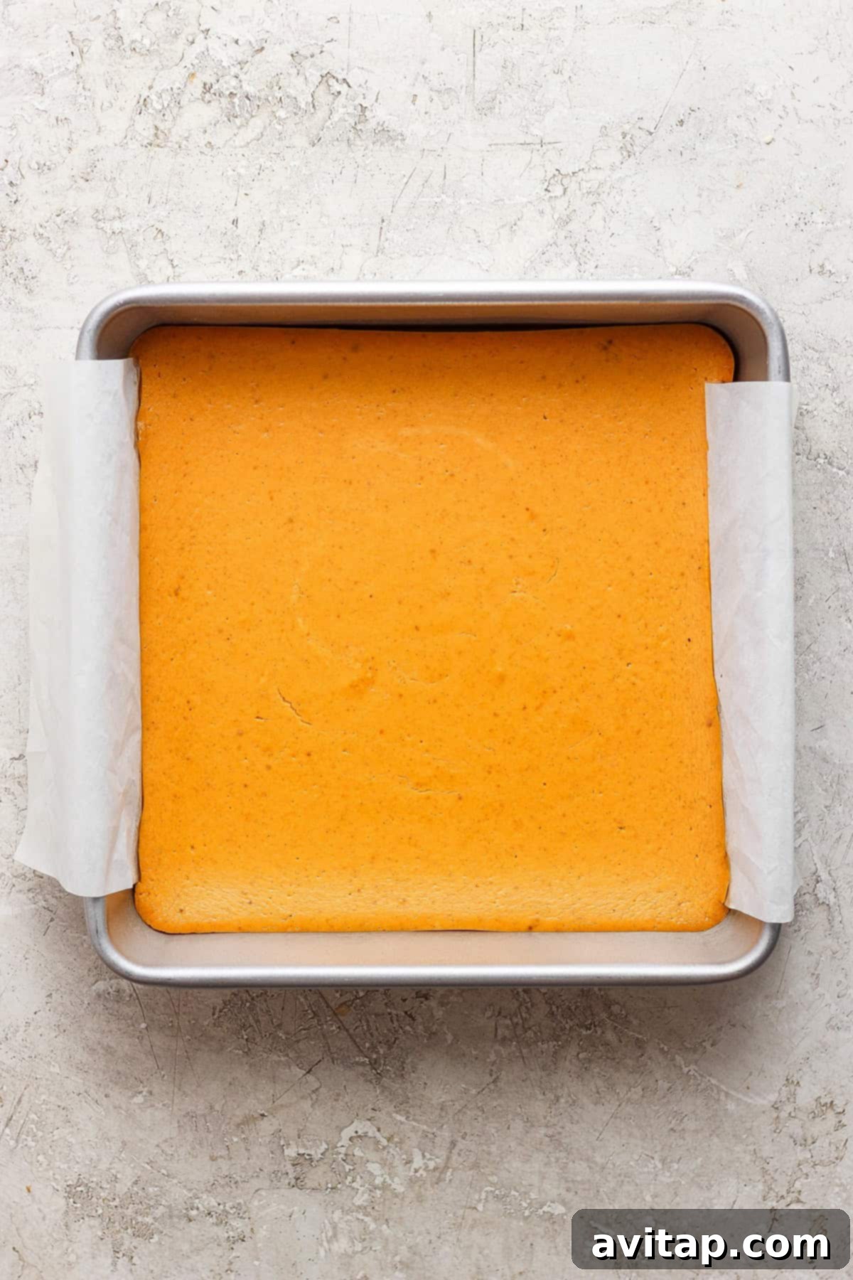 A freshly baked square pumpkin bar, a beautiful golden-brown, cooling in a parchment-lined baking pan on a light, textured surface, ready to be sliced and served.