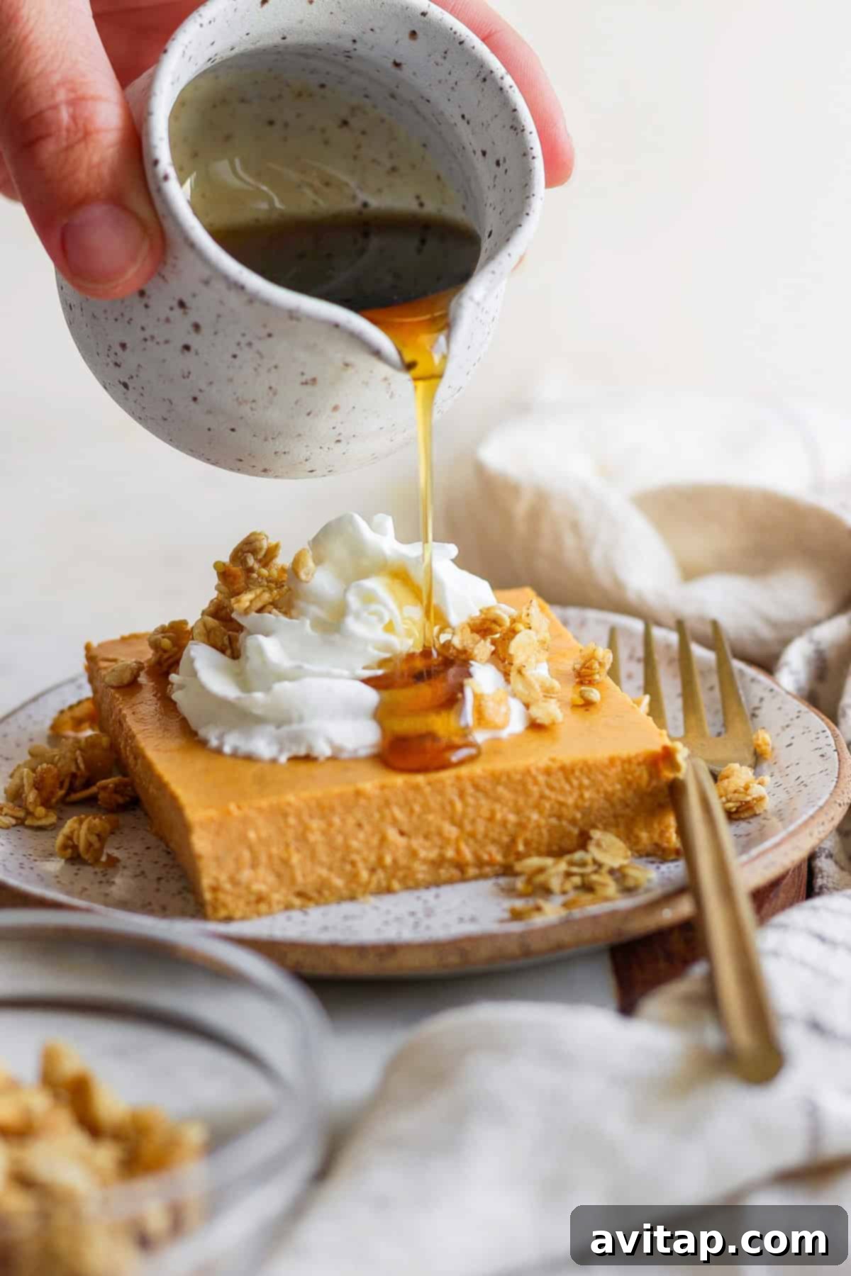 A close-up shot of a hand drizzling glistening maple syrup over a beautifully plated slice of high-protein pumpkin bar, adorned with whipped cream and crunchy granola. A fork and napkin are neatly arranged beside it, ready for enjoyment.