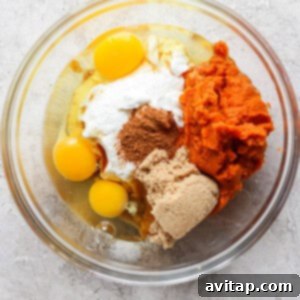 A glass mixing bowl displays raw ingredients including eggs, plain yogurt, vibrant pumpkin puree, light brown sugar, and aromatic spices, all prepared and ready for a thorough whisking.