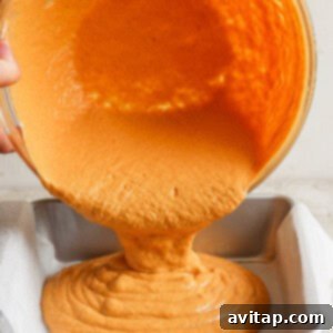 A pair of hands gently pours the vibrant orange batter from a mixing bowl into a square baking pan that is lined with parchment paper, ensuring even distribution for baking.