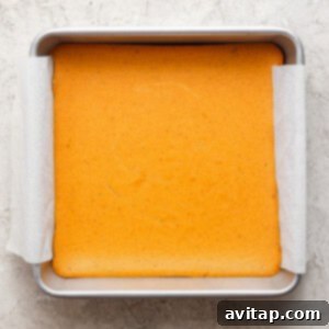 A square baking pan, lined with parchment paper, holds a freshly baked cake with a light golden-brown top, cooling on a textured, light-colored kitchen surface, awaiting slicing.