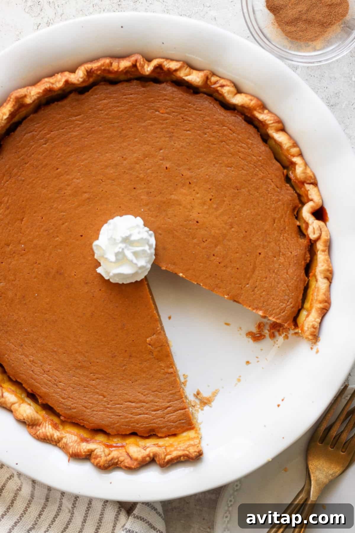 Perfectly Spiced Pumpkin Pie 4 A pumpkin pie with a slice missing, topped with a dollop of whipped cream, in a round white pie dish. There is a folded towel and two gold forks next to the dish.