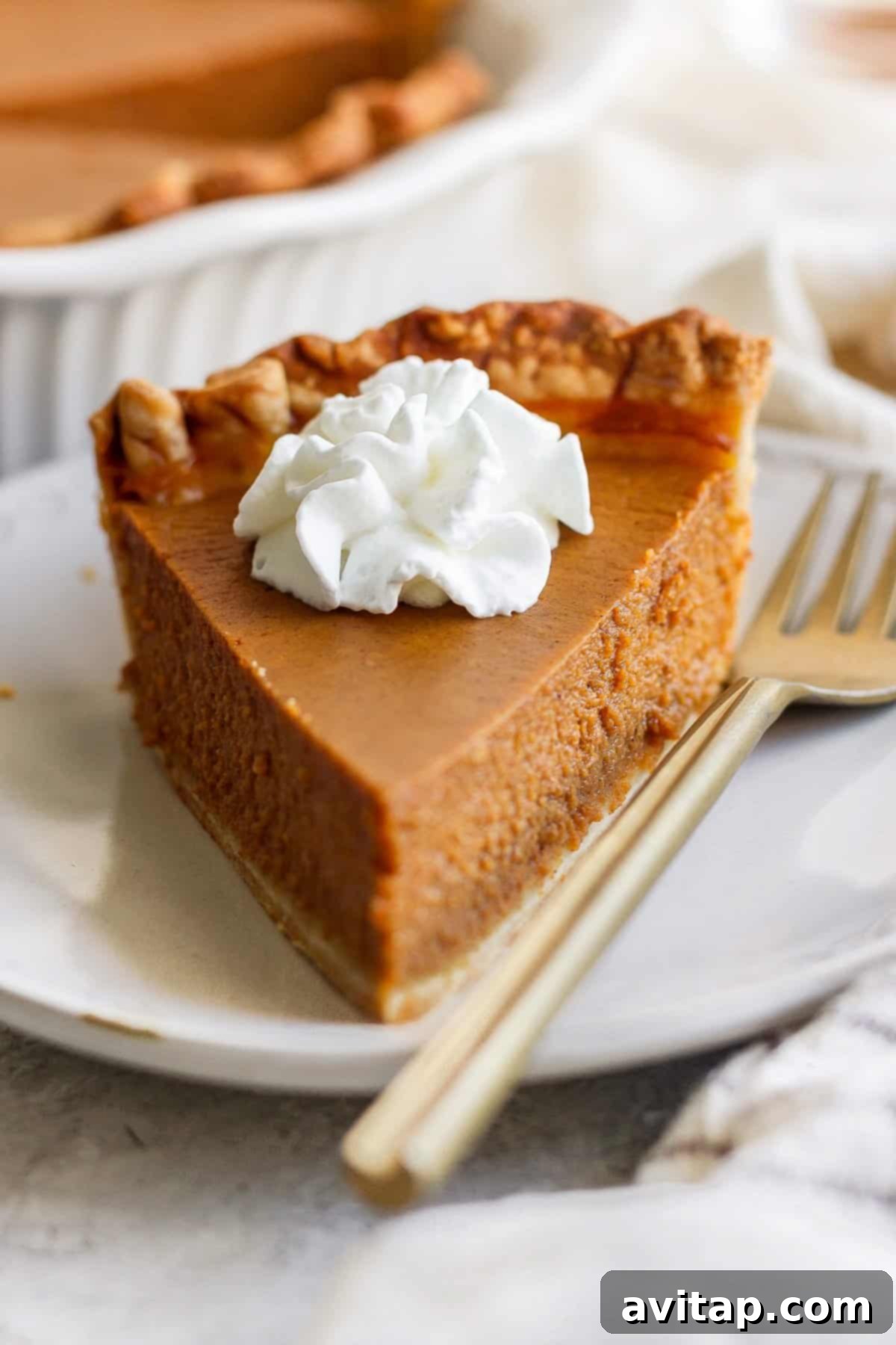 Perfectly Spiced Pumpkin Pie 5 A slice of pumpkin pie with a dollop of whipped cream on top, placed on a white plate next to a fork.
