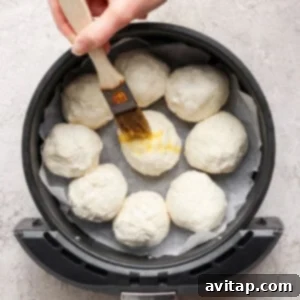 A hand holds a pastry brush, applying a shiny egg wash to the tops of dough balls arranged in an air fryer basket, which is lined with parchment paper.