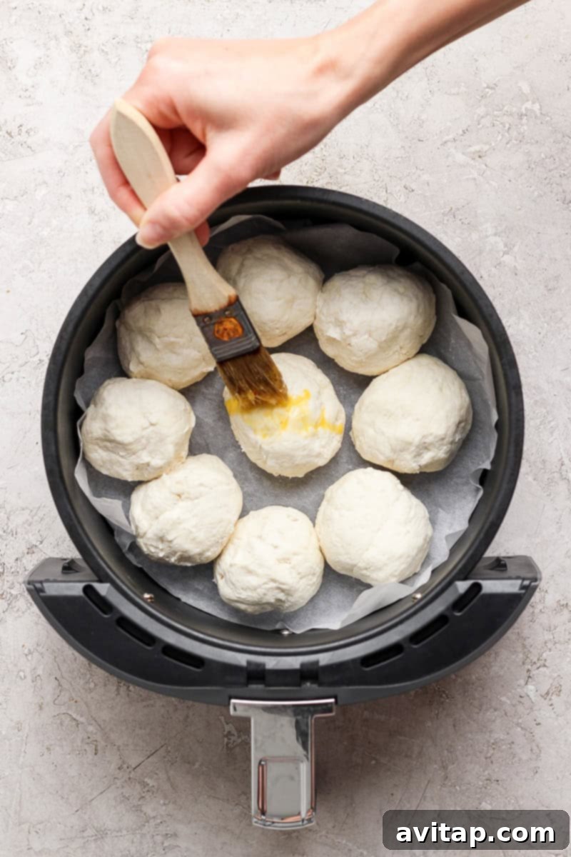 A hand expertly brushes egg wash onto uncooked dough balls, neatly arranged in an air fryer basket lined with parchment paper.