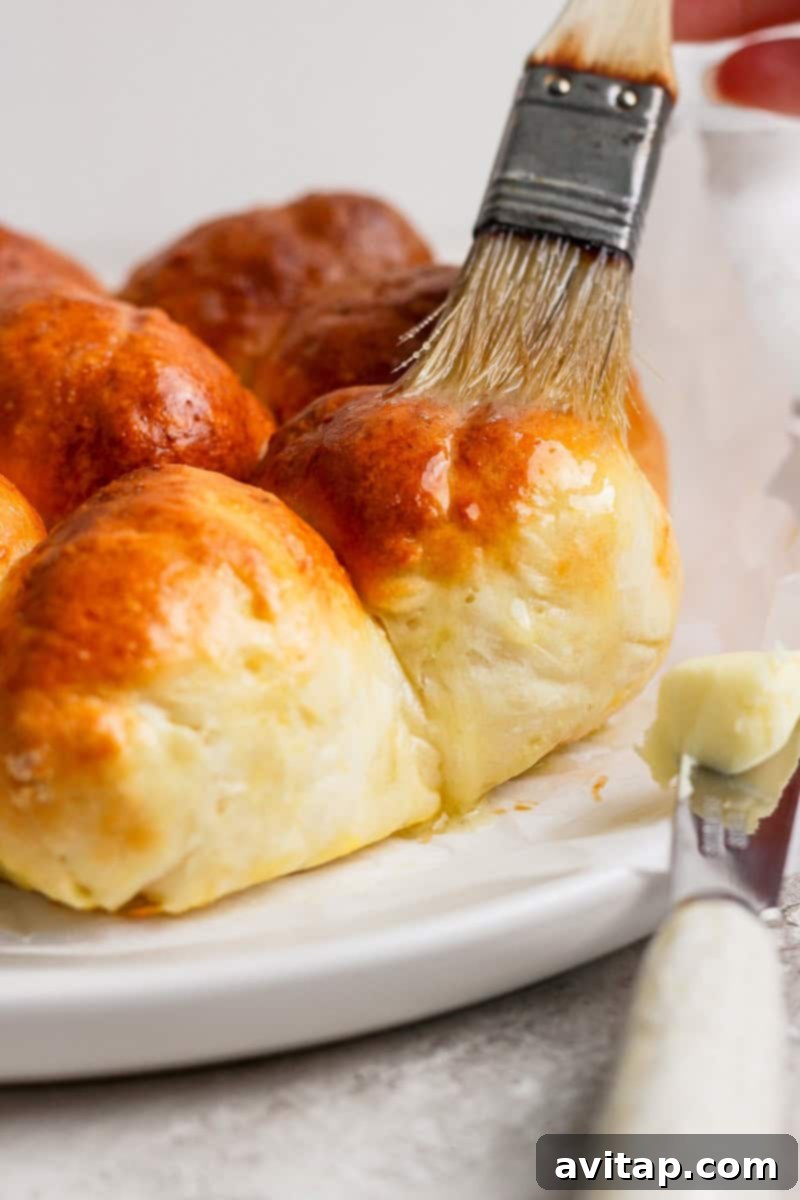 A pastry brush applies melted butter to freshly baked, golden-brown bread rolls arranged on a pristine white plate.