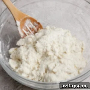 A wooden spoon rests in a glass bowl filled with a mixture of flour and a white ingredient, likely dough, on a textured countertop, ready for mixing.