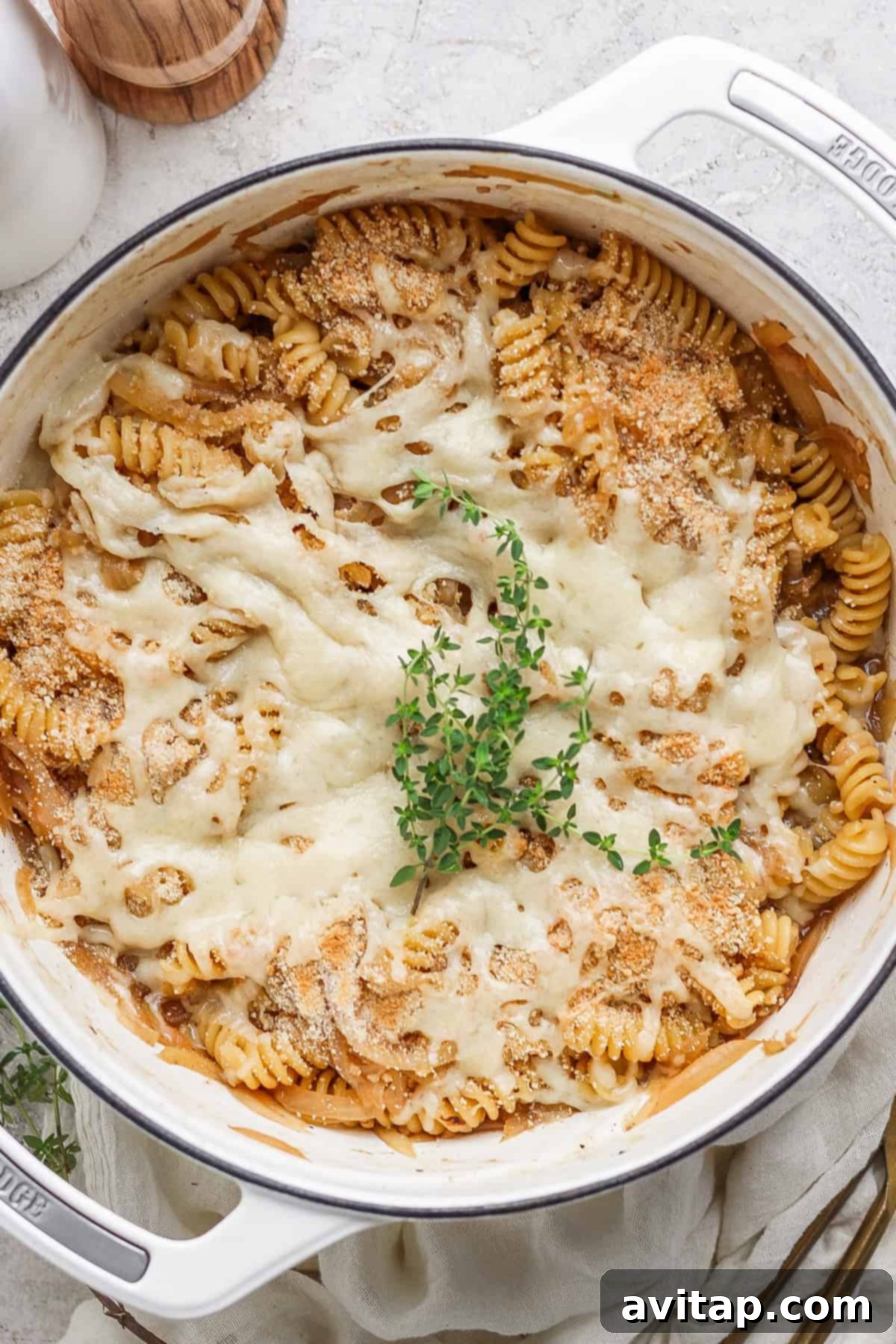 Sweet and Savory Onion Pasta 2 A baked pasta dish in a white pot, featuring rotini pasta covered with melted cheese and garnished with a sprig of fresh herbs on top.