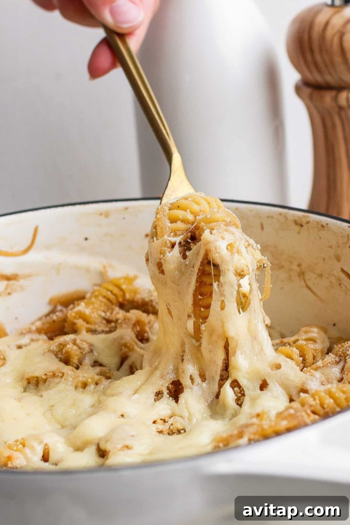 Sweet and Savory Onion Pasta 7 A hand using a golden fork to lift cheesy pasta from a white pot, with melted cheese stretching from the pot.