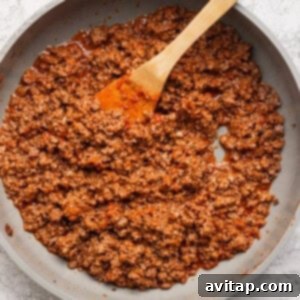 Cooked ground beef in a light-colored skillet with a wooden spatula, ready to be combined with the sauce, placed on a textured white surface.