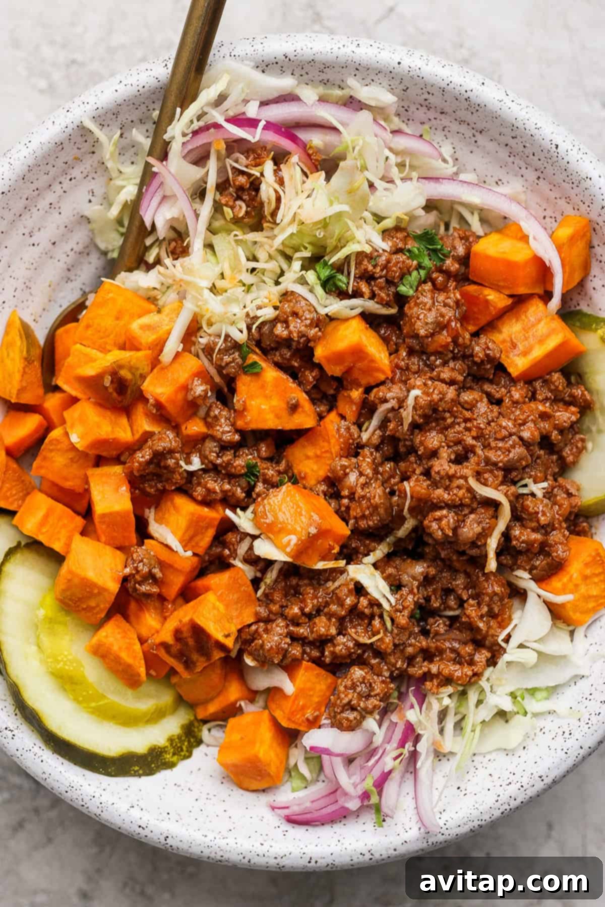 A meticulously arranged Sloppy Joe Bowl featuring seasoned ground beef, vibrant diced sweet potatoes, crunchy pickle slices, fresh shredded lettuce, and delicate red onion rings, accompanied by a fork for immediate enjoyment.