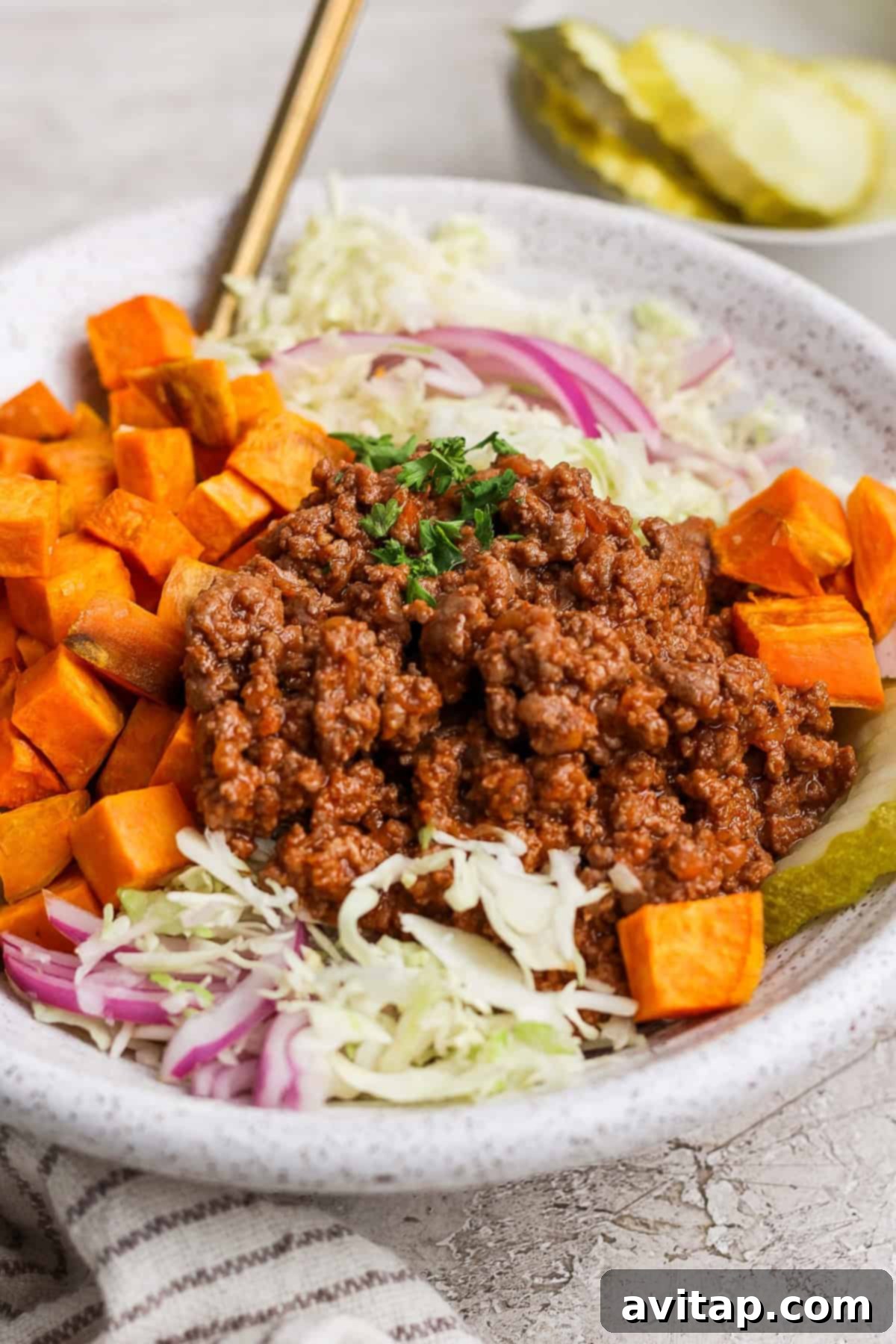 A beautifully composed Sloppy Joe Bowl with a base of shredded cabbage and red onion, topped with tender diced sweet potatoes, a generous scoop of savory seasoned ground meat, and garnished with fresh parsley. Tangy pickle slices are artfully arranged in the background.