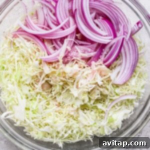 A clear glass bowl containing a vibrant mixture of sliced red onions and shredded cabbage, ready for dressing, placed on a light gray surface.