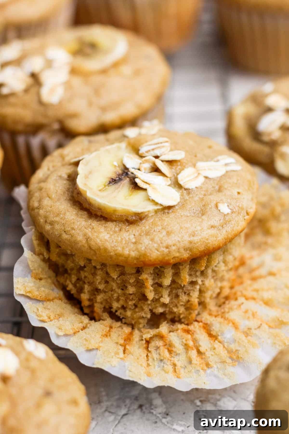 A perfectly baked banana oatmeal muffin, topped with a banana slice and a sprinkle of oats, partially unwrapped on a rustic wire cooling rack, promising a wholesome and delicious bite.