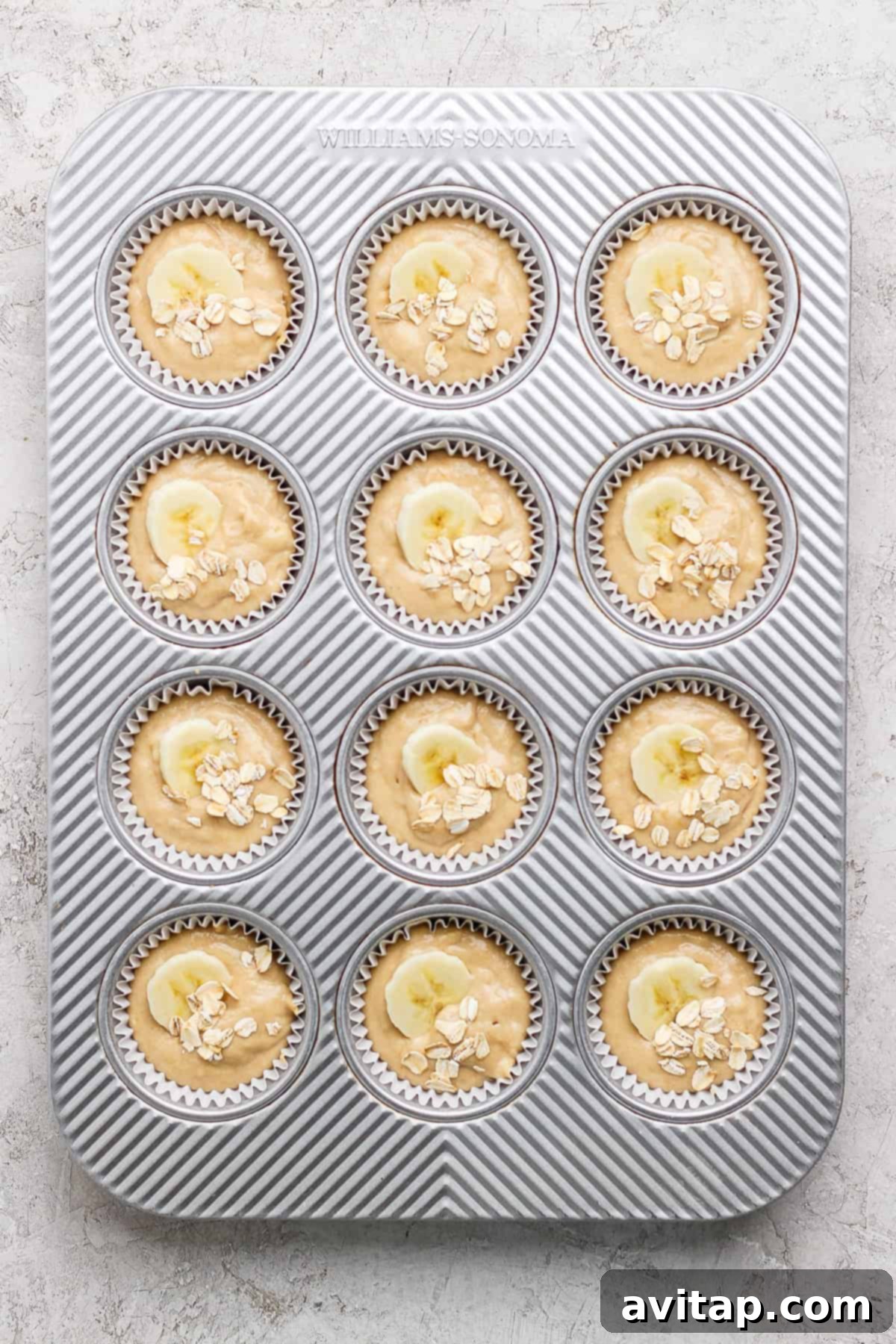 A muffin tray carefully filled with twelve portions of banana oatmeal muffin batter, each beautifully adorned with thin banana slices and a sprinkle of oats, poised for baking.