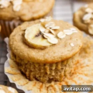 A banana muffin topped with a banana slice and oats, with a paper liner partially peeled back, showcasing its moist texture.