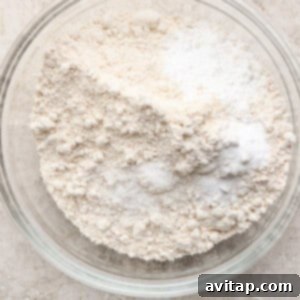A clear glass bowl containing a mixture of oat flour, baking soda, baking powder, and salt, ready to be whisked together on a textured beige surface.