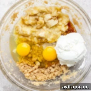 A glass bowl containing mashed bananas, two eggs, Greek yogurt, brown sugar, and melted coconut oil, ready to be mixed into a creamy wet batter.