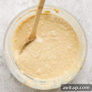 A glass bowl filled with creamy banana oatmeal muffin batter with a wooden spoon resting in it, showcasing the perfectly mixed consistency on a textured white surface.