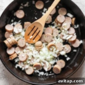 A black skillet containing sliced sausages and diced onions being stirred with a wooden spoon.