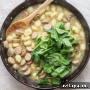 A skillet containing gnocchi, sliced sausages, and fresh spinach, with a wooden spoon resting inside.