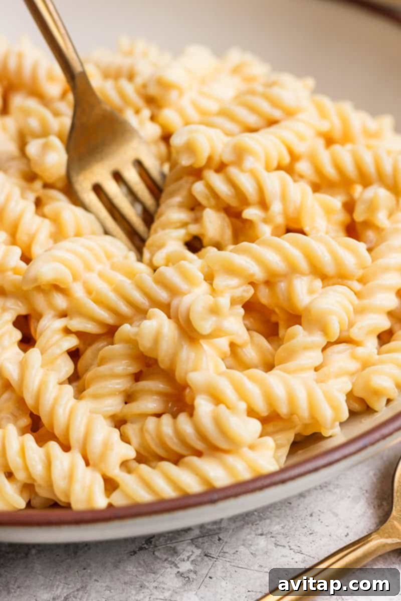 A fork twirls creamy cheese sauce-covered rotini pasta in a bowl on a textured surface, showcasing the rich, glossy sauce.