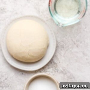 Dough ball on a plate, a measuring cup with water, and a bowl of sugar on a textured surface.