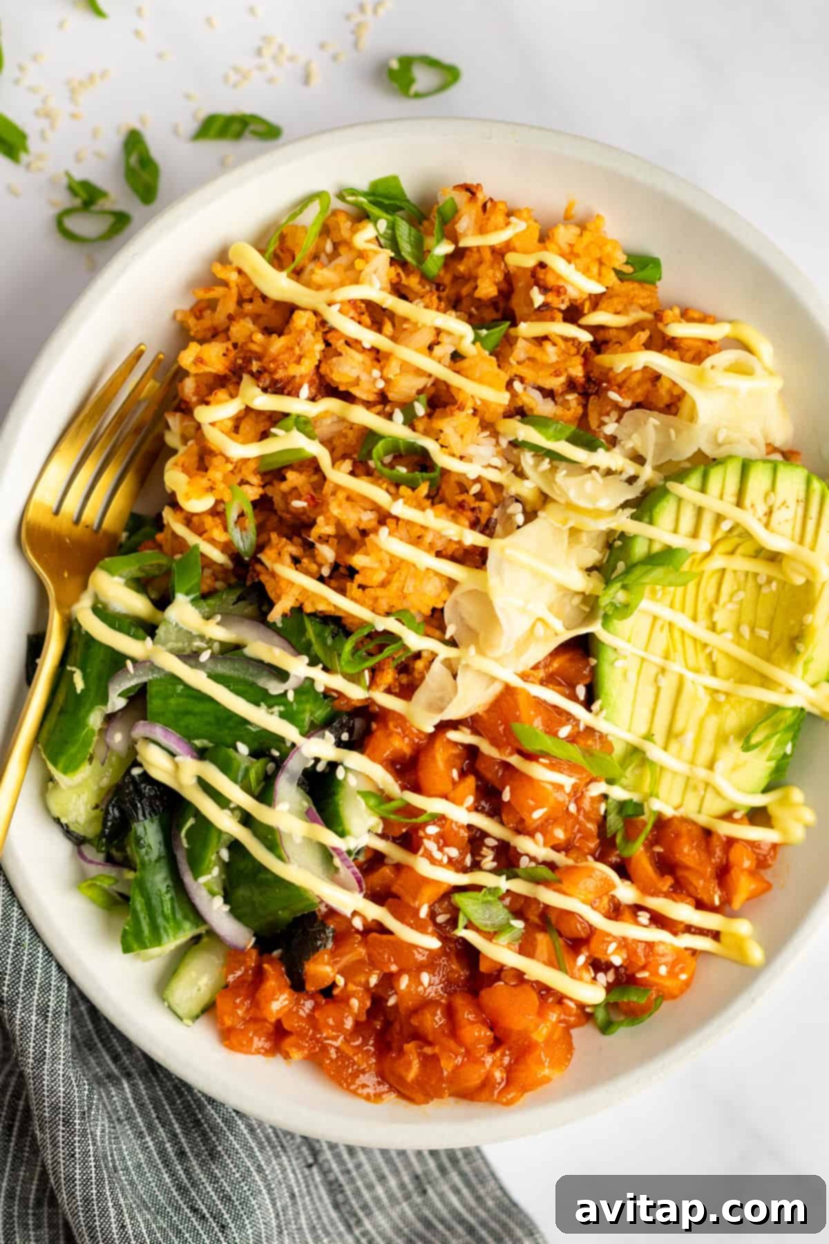 A bowl of rice topped with avocado slices, marinated raw fish, sliced vegetables, shredded cabbage, and drizzled with a creamy sauce. A gold fork rests on the side.