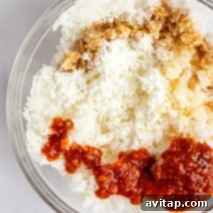 A bowl containing white rice, crumbled cheese, soy sauce, and red chili sauce on a light surface.
