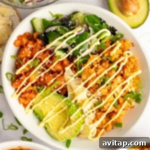 A bowl of rice topped with avocado, kimchi, salad greens, drizzled with mayonnaise, next to half an avocado and additional side dishes.
