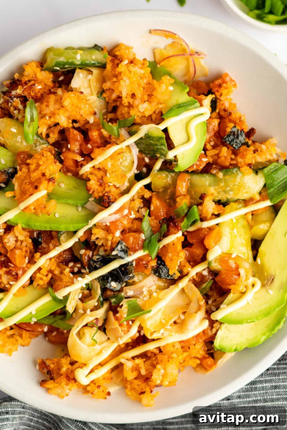 A plate of seasoned rice topped with sliced avocado, cucumber, seaweed, and a drizzle of cream sauce.