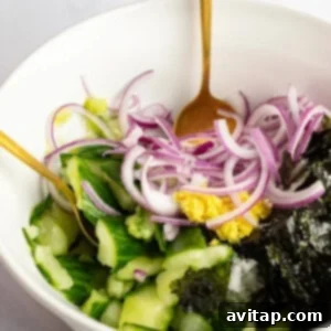 A hand mixes a salad in a white bowl with a gold spoon. The salad contains sliced cucumbers, red onion, seaweed, and a dollop of mustard.