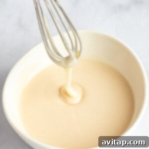 Fit Frosted Pastries 14 A whisk with creamy batter drips into a white bowl on a light surface, illustrating the process of making the glaze.
