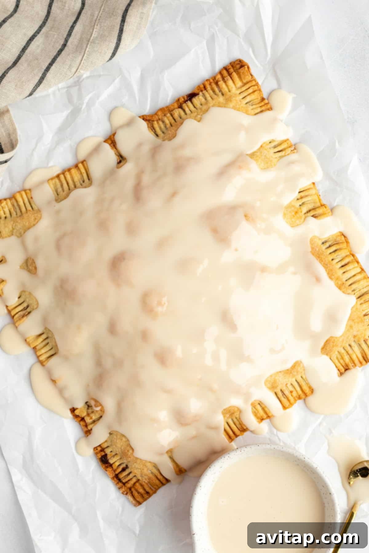 Fit Frosted Pastries 6 A large rectangular pastry with perfectly ridged edges, beautifully covered in a smooth white icing, placed on crinkled parchment paper next to a small bowl of extra icing, ready to be portioned and enjoyed.