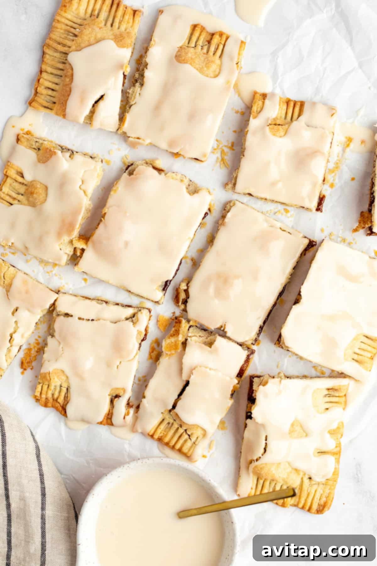 Fit Frosted Pastries 7 Nine perfectly iced rectangular pastries with a delightful crumbly texture, meticulously arranged on parchment paper. A small bowl of extra icing with a spoon sits at the bottom left, ready for any additional touches.