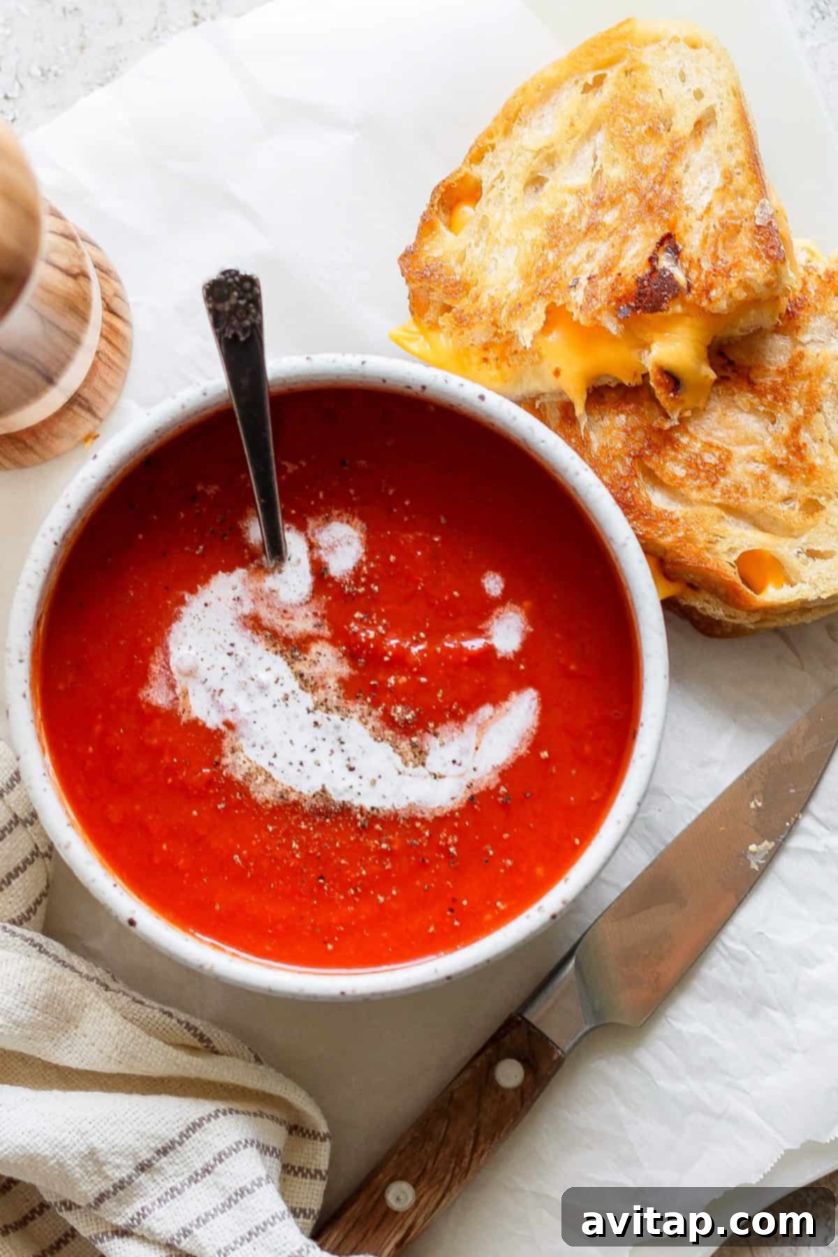 A steaming bowl of creamy tomato soup, artfully swirled with a hint of fresh cream and sprinkled with cracked black pepper. Beside it rests a golden-brown, crispy grilled cheese sandwich, cut diagonally, revealing its gooey, melted interior. A gleaming knife and a striped fabric napkin complete this inviting and classic comfort food scene.