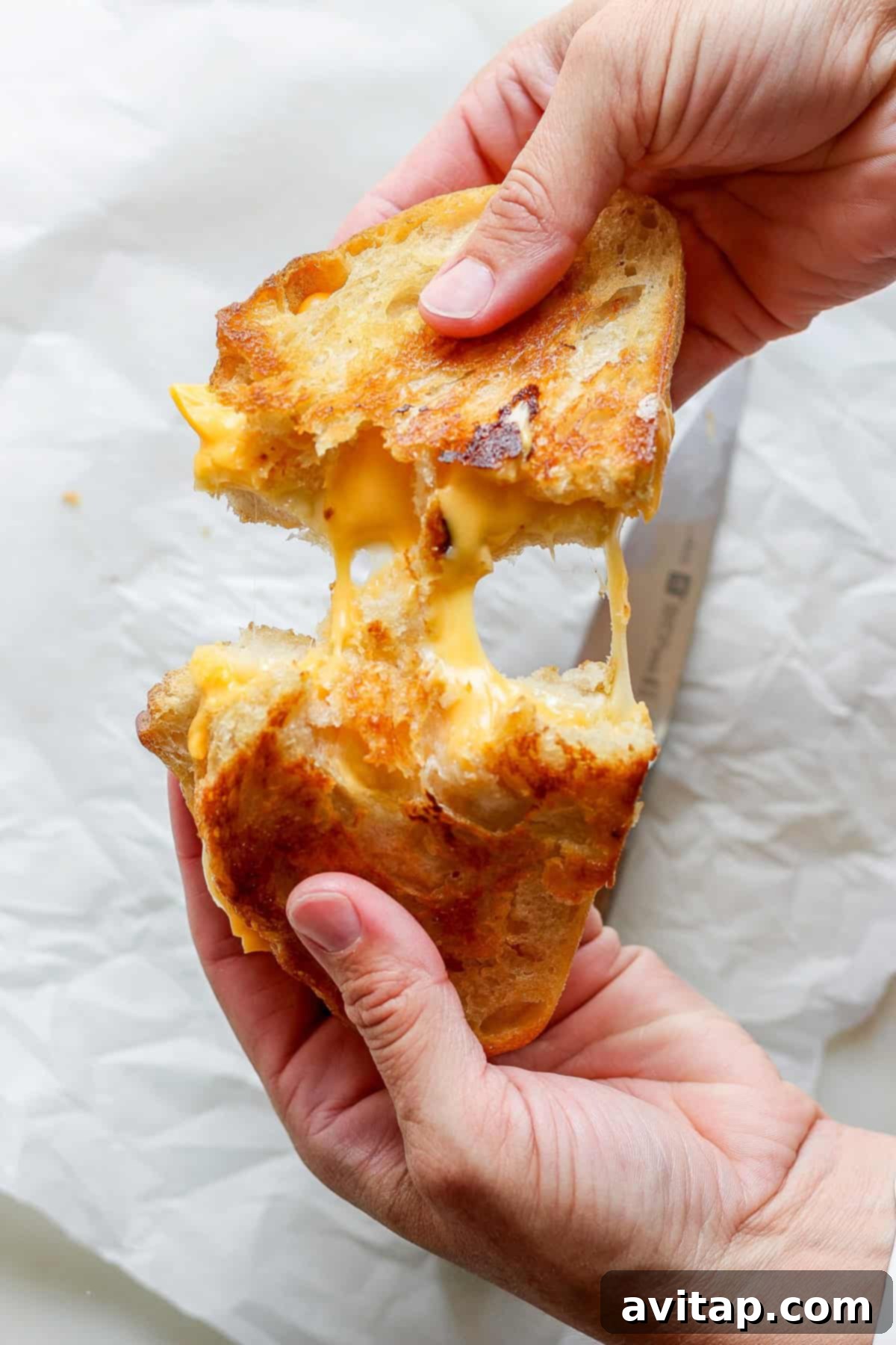 Hands gently pulling apart a perfectly golden grilled cheese sandwich, revealing luscious strands of melted cheese stretching invitingly between the toasted sourdough slices. This iconic image highlights the gooey perfection that pairs exquisitely with a warm bowl of tomato soup.