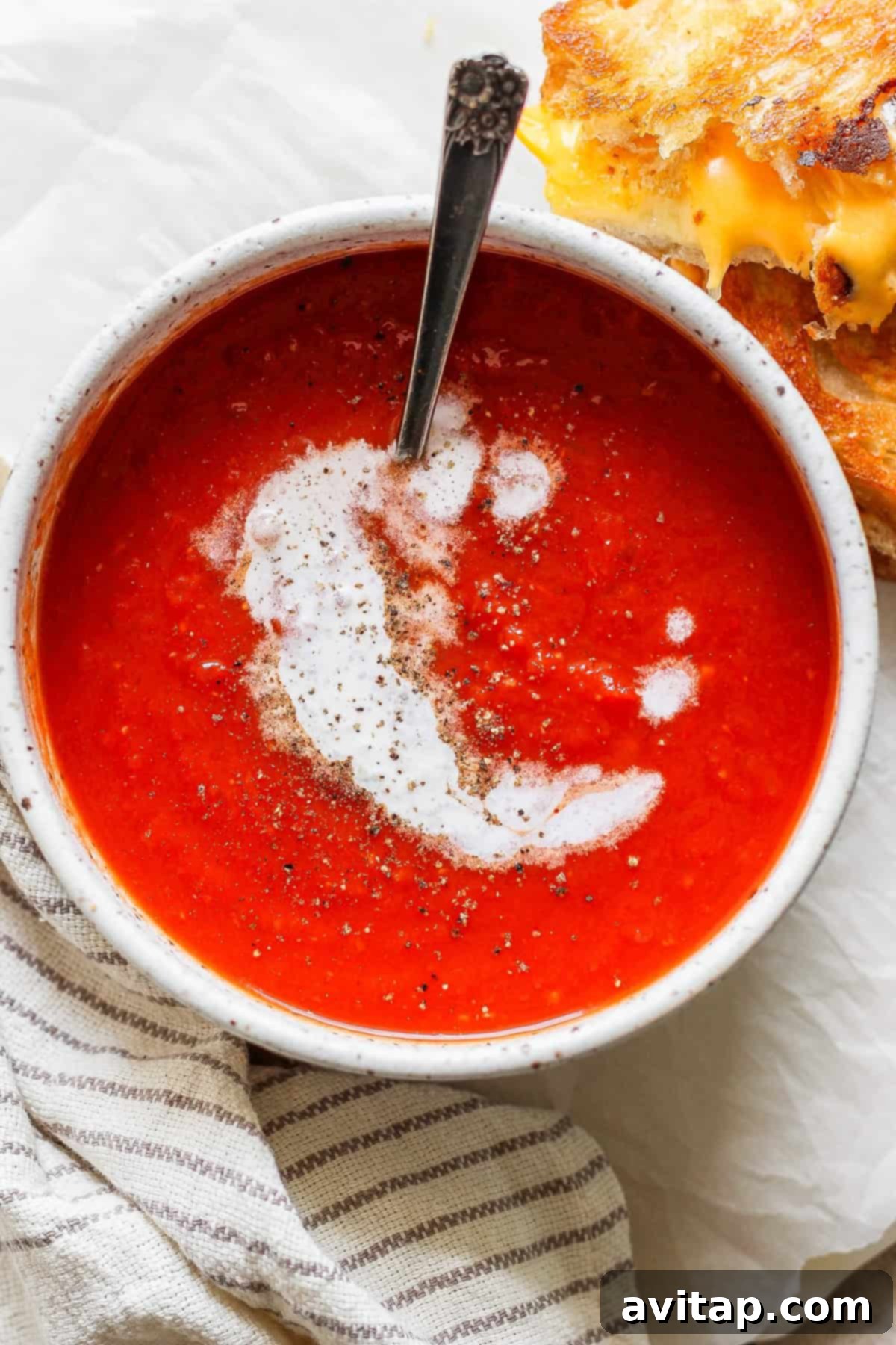 A beautifully presented bowl of creamy tomato soup, elegantly swirled with cream and dusted with pepper, creating an inviting visual. Beside it, a perfectly toasted grilled cheese sandwich, golden and crisp, offers a delectable contrast. A spoon rests invitingly in the bowl, hinting at the delightful experience to come, while a neatly folded striped cloth napkin completes this comforting and classic culinary scene.