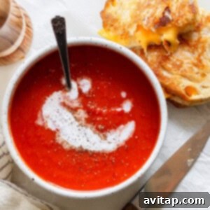 A bowl of rich tomato soup swirled with cream and pepper sits beside a perfect pairing: a crispy grilled cheese sandwich. A knife rests nearby on the table, alongside a neatly folded napkin, completing the classic duo's delicious presentation.