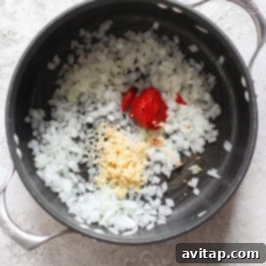 Chopped onions, minced garlic, and dollops of tomato paste in a black pot on a light textured surface evoke the comforting aroma of grilled cheese and tomato soup.