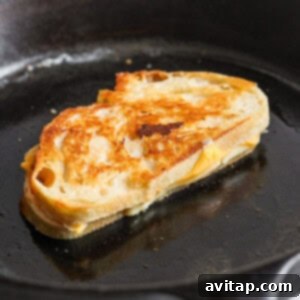 A golden-brown grilled cheese sandwich sizzles enticingly in a black skillet, its edges crispy and its cheese visibly melting. This perfect grilled cheese is an ideal partner for a comforting bowl of tomato soup.