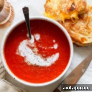 A bowl of rich, red tomato soup, artfully swirled with a touch of cream and topped with freshly ground pepper. Beside it, a perfectly toasted grilled cheese sandwich, cut in half, sits invitingly. A knife and a striped napkin complete this iconic comfort food duo.